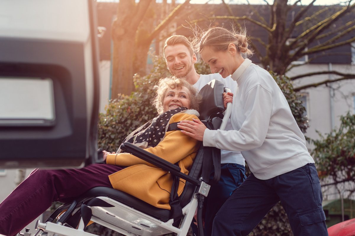 Two helpers picking up disabled senior woman in wheelchair for transport